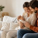 Jeunes parents assis sur un canapé avec leur nouveau-né dans un salon lumineux, illustrant le congé supplémentaire de naissance et le temps passé en famille après l’arrivée d’un enfant.