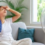 Une femme assise sur un canapé clair semble souffrir de la chaleur, tenant un éventail tout en essuyant son front dans un geste de fatigue et d’inconfort.